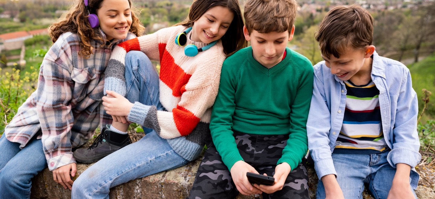 2 Mädchen und 2 Jungen sitzen auf einer Mauer und schauen auf das Smartphone des einen Jungen.